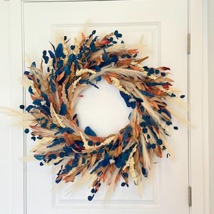 May include: A decorative wreath with a circular design, featuring a mix of dried and faux foliage. The wreath incorporates elements in shades of blue, orange, beige, and cream, creating a natural and textured appearance. It is displayed on a white door.