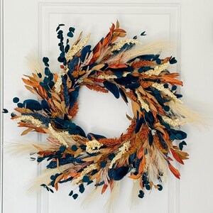 May include: A fall-themed wreath with a variety of dried flowers and foliage. The wreath features a mix of colors, including orange, navy blue, cream, and beige. The wreath is circular and is displayed on a white door.