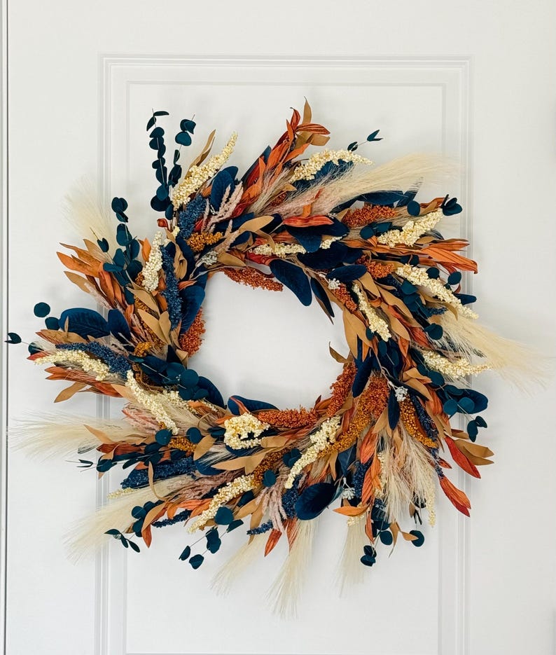 May include: A fall-themed wreath with a variety of dried flowers and foliage. The wreath features a mix of colors, including orange, navy blue, cream, and beige. The wreath is circular and is displayed on a white door.