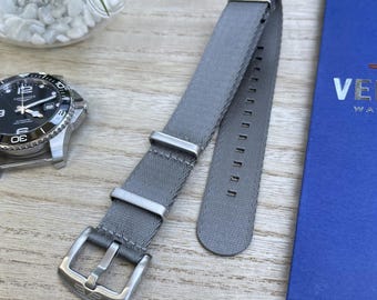 May include: A gray nylon watch strap with stainless steel hardware. The strap is laid out on a wooden surface next to a watch with a black face and a blue box with the word "VELA" on it. The watch strap is designed to be durable and stylish.