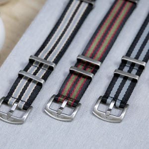Peut inclure: Trois bracelets de montre avec boucles en acier inoxydable. Un bracelet est noir avec des rayures grises, un autre est noir avec des rayures rouges et vertes, et le troisième est noir avec des rayures grises. Une montre avec un cadran noir est partiellement visible.