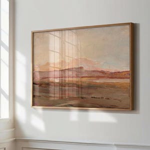 May include: A framed landscape painting with a warm color palette. The artwork depicts a mountain range and a body of water, with hues of brown, pink, and beige. The frame is a light brown color. The painting is hanging on a white wall.