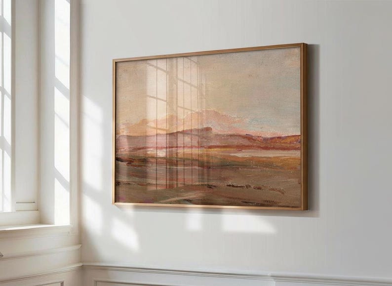 May include: A framed landscape painting with a warm color palette. The artwork depicts a mountain range and a body of water, with hues of brown, pink, and beige. The frame is a light brown color. The painting is hanging on a white wall.