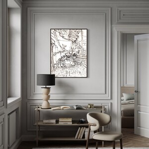 May include: A black and white line drawing of a street scene with people sitting at tables outside a cafe. The drawing is framed and hanging on a wall in a room with a wooden floor, a white chair, and a bookshelf.