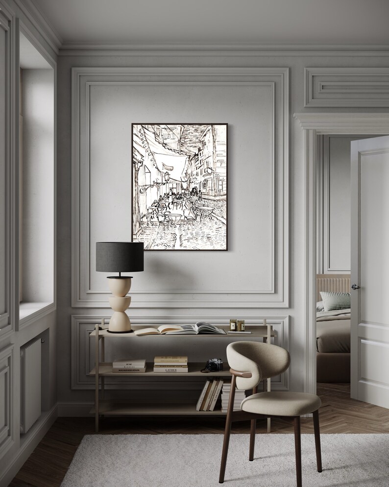 May include: A black and white line drawing of a street scene with people sitting at tables outside a cafe. The drawing is framed and hanging on a wall in a room with a wooden floor, a white chair, and a bookshelf.
