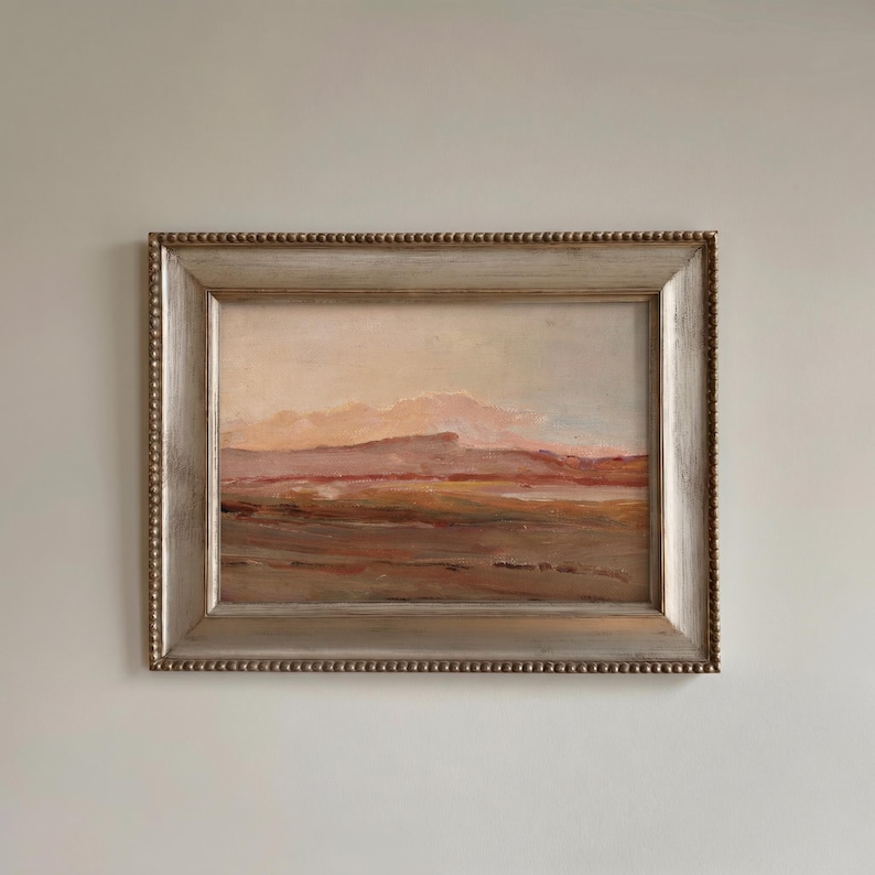 May include: A framed landscape painting with a silver-colored beaded frame. The artwork depicts a muted color palette of pink, brown, and beige, suggesting a sunset or sunrise over a natural scene. The painting is rectangular.
