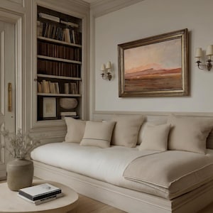 May include: A cozy living room scene featuring a cream-colored sofa with several pillows, a round coffee table, and a framed landscape painting. A built-in bookcase is visible in the background, filled with books. The room is decorated in neutral tones.