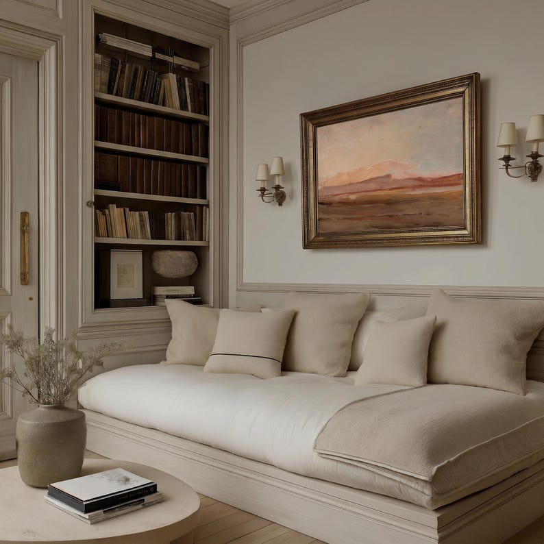 May include: A cozy living room scene featuring a cream-colored sofa with several pillows, a round coffee table, and a framed landscape painting. A built-in bookcase is visible in the background, filled with books. The room is decorated in neutral tones.