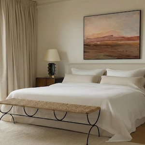 May include: A bedroom scene featuring a large bed with white linens, pillows, and a neutral-toned headboard. A woven bench sits at the foot of the bed. The room is decorated with a landscape painting and two lamps.