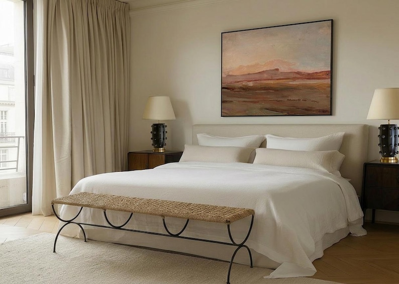 May include: A bedroom scene featuring a large bed with white linens, pillows, and a neutral-toned headboard. A woven bench sits at the foot of the bed. The room is decorated with a landscape painting and two lamps.