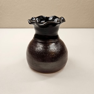 May include: A small, dark brown ceramic vase with a black, ruffled rim.