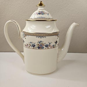 May include: A white ceramic coffee pot with a gold trim and floral design. The lid has a gold knob and a floral design.