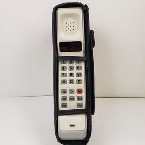 May include: A vintage off-white portable cellular phone encased in a black leather carrying sleeve. The device features a numeric keypad, function buttons labeled Rcl, Clr, Snd, Sto, Lock, End, Pwr, and Vol, and the BellSouth Mobility logo.