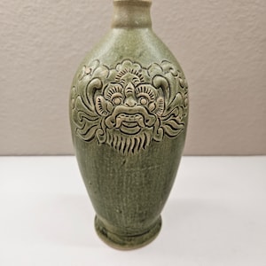 May include: A green ceramic vase with a raised design of a mythical creature's face. The vase has a rounded body and a narrow neck.