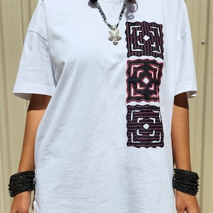 May include: A white short-sleeved t-shirt featuring three vertical black and red square embroidered patches on the right side. The shirt is paired with a beaded necklace with a silver pendant and stacks of thin black bracelets on both wrists.