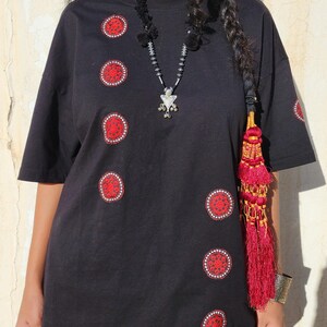 May include: A black short-sleeved t-shirt featuring a series of circular red and white embroidered patches arranged in an asymmetrical pattern across the front and on the sleeve. The shirt has a crew neckline and a loose, relaxed fit silhouette.