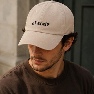 May include: A beige baseball cap featuring the embroidered black text "¿Y si sí?" on the front panel. The cap has a structured crown, a curved brim, and stitched eyelets for ventilation, providing a classic look with a simple, bold statement.