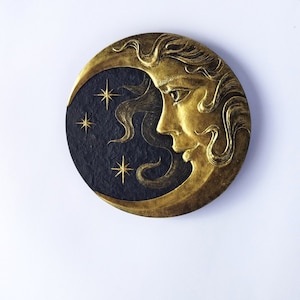 Può includere: Un decoro da parete a forma di mezzaluna dorata e nera con il viso di una donna di profilo. La luna ha una superficie testurizzata ed è decorata con stelle dorate.