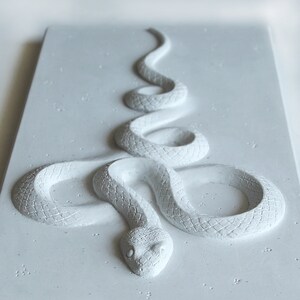 Op de afbeelding: Een wit, getextureerd, slangensculptuur in een opgerolde positie. De slang is gemaakt van een materiaal dat lijkt op beton of gips.