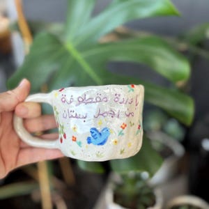 Puede incluir: Taza de cerámica blanca con un pájaro azul y diseños florales. La taza tiene el texto "لا تزين مقطوعة من اجمل سنين" en árabe.
