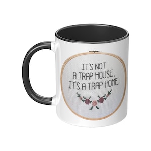 May include: White ceramic mug with a black rim and handle. The mug features a cross-stitch design with the text "It's not a trap house, it's a trap home."