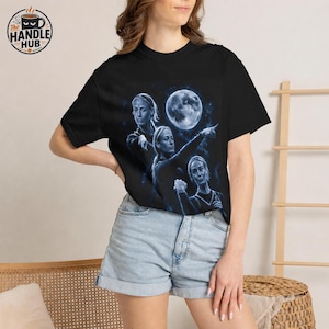 May include: A black short-sleeve t-shirt featuring a blue-toned graphic print of three identical figures pointing toward a large, glowing full moon. The design includes starry accents and a celestial theme, set against a dark background on the chest.