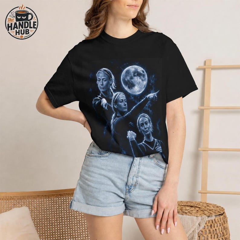 May include: A black short-sleeve t-shirt featuring a blue-toned graphic print of three identical figures pointing toward a large, glowing full moon. The design includes starry accents and a celestial theme, set against a dark background on the chest.