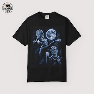 May include: A black short-sleeve t-shirt featuring a blue-toned graphic print of three figures in various poses beneath a full moon. The design includes a circular logo in the top left corner with the text The Handle Hub and a coffee cup icon.
