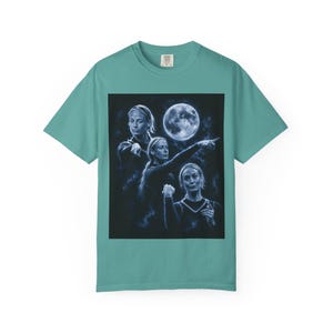 May include: A teal short-sleeve t-shirt featuring a graphic print of a person in three different poses against a dark background with a full moon. The person is shown pointing and gesturing in a blue-toned, celestial-themed artistic composition.