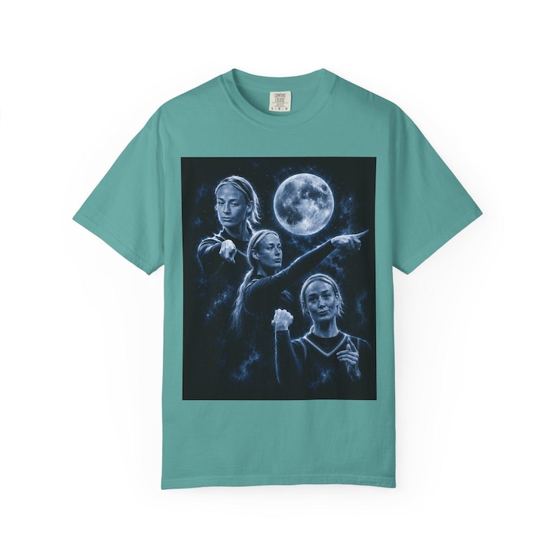 May include: A teal short-sleeve t-shirt featuring a graphic print of a person in three different poses against a dark background with a full moon. The person is shown pointing and gesturing in a blue-toned, celestial-themed artistic composition.