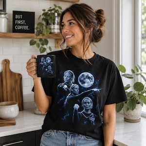 May include: A black t-shirt featuring a blue-toned graphic of a person in three different poses, including one pointing, set against a full moon and starry background. The same graphic is printed on a black ceramic mug held in the hand.