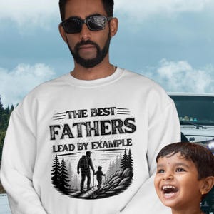 Peut inclure: Sweat-shirt blanc Gildan 18000 avec le texte "THE BEST FATHERS LEAD BY EXAMPLE" et un graphique d'un père et d'un enfant en randonnée. L'image montre un homme et un enfant devant un véhicule et un fond de montagne.