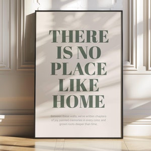 May include: A beige poster with black framing, featuring the text "THERE IS NO PLACE LIKE HOME" in dark green. The text is in a bold, sans-serif font. Below the text, a smaller font reads "Between these walls, we've written chapters of joy, painted memories in every colour, and grown roots deeper than time."