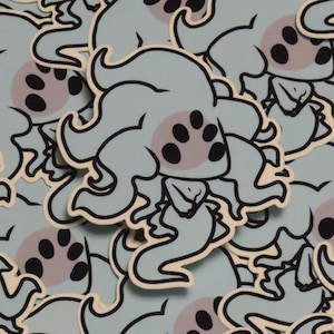 May include: A close-up of a pile of blue and black stickers featuring a cartoon creature with multiple tentacles and large eyes. The creature has a light blue body with black eyes and a pink spot on its head.