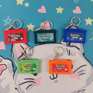 May include: A collection of six colorful Pokémon game cartridge keychains. The keychains are red, green, blue, orange, and green. Each keychain features the game's title, such as "Ruby Version" and "LeafGreen Version."