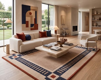 May include: A large rectangular area rug featuring a geometric border in navy blue, cream, and rust orange. The rug is placed in a living room with a cream sofa, a low-profile stone coffee table, and a white armchair on light wood flooring.