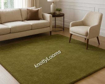May include: A large, olive green rectangular rug dominates a living room setting. The rug is placed on a hardwood floor, beneath a beige sofa, armchair, and a small side table with a lamp. The text "knottyLooms" is visible on the rug.