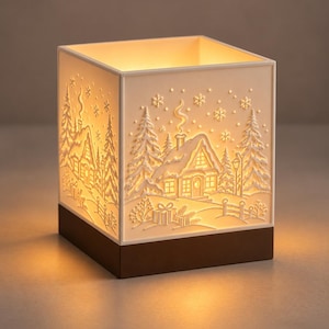May include: A square white luminary with a dark brown base, featuring an embossed winter scene of a snow-covered cottage, pine trees, and falling snowflakes. The interior is illuminated with a warm glow, highlighting the relief details.