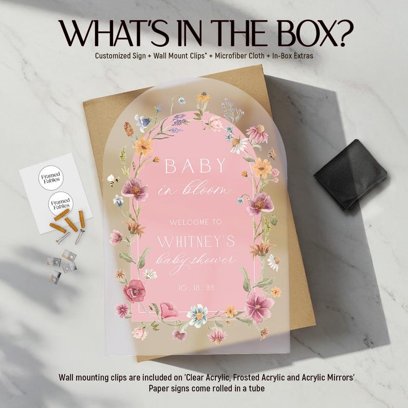 Op de afbeelding: Een babyborrelbord met een roze achtergrond en bloemenrand. Op het bord staat "BABY in bloom" en verwelkomt gasten bij "Whitney's baby shower" op 15 oktober 2025. De afbeelding bevat ook wandmontageclips en een microvezeldoek.