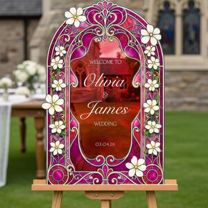 May include: An arched wedding welcome sign featuring a vibrant magenta and gold stained glass border adorned with white flowers. The centre displays the text: WELCOME TO Olivia & James WEDDING 03.04.26, set against a blurred stone building background.