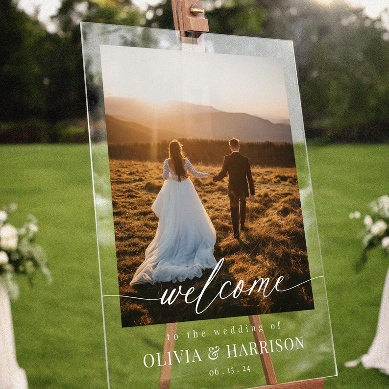 May include: A wedding welcome sign with a photo of a couple walking away from the camera in a field. The sign says "welcome to the wedding of Olivia & Harrison 06.15.24".