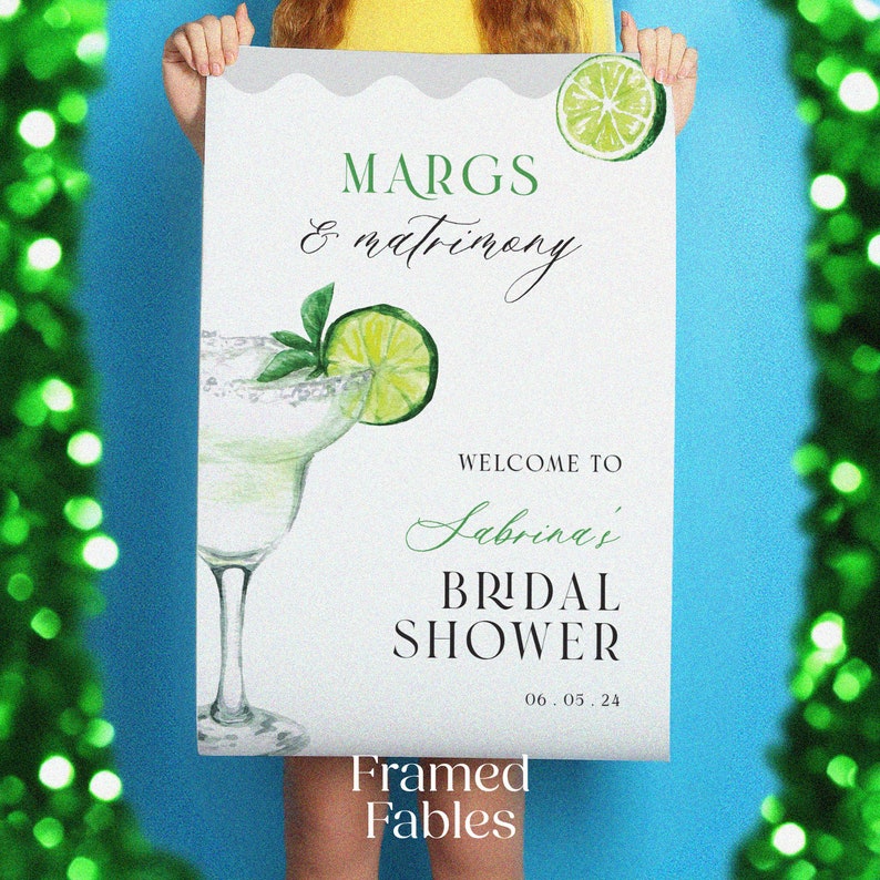 May include: A white sign with green and white watercolor illustrations of a margarita and lime wedges. The text on the sign reads "MARGS & matrimony WELCOME TO Sabrina's BRIDAL SHOWER 06.05.24 Framed Fables".