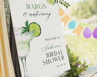 May include: A white sign with green text and a watercolor illustration of a margarita glass with a lime wedge. The text reads "Margs & Matrimony" and "Welcome to Sabrina's Bridal Shower 06.03.24".