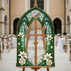 K&ouml;nnte beinhalten: Ein bogenf&ouml;rmiges Hochzeitsschild mit einem gr&uuml;nen Buntglasrahmen und Illustrationen von wei&szlig;en Lilien und Rosen. Der mittlere Text lautet: Welcome TO THE WEDDING OF KRYSTAL and WILLIAM NEW YORK, USA 06.08.26, mit den Initialen K/W im oberen Kreis.