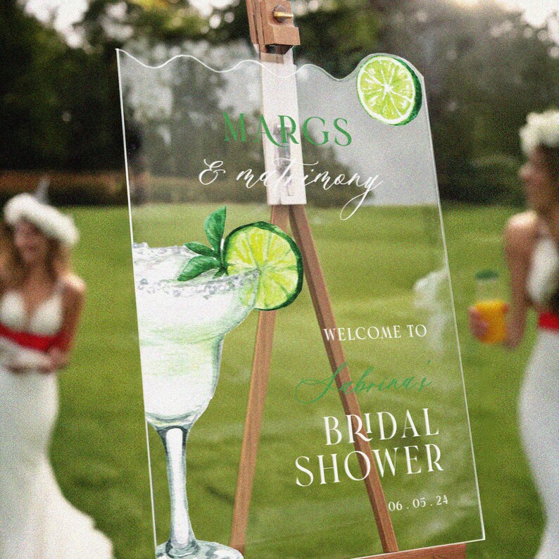 May include: A clear acrylic sign with a white background and green text. The sign says "MARGS & matrimony" and "WELCOME TO Sabrina's BRIDAL SHOWER 06.05.24". There is a green lime wedge and a margarita glass with a lime wedge on the sign.