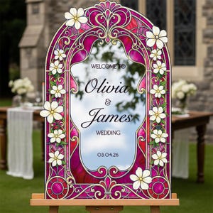 May include: An arched wedding sign featuring a vibrant magenta and gold stained glass border with white floral accents. The centre displays the text: WELCOME TO Olivia & James WEDDING 03.04.26. The sign is mounted on a wooden easel stand.