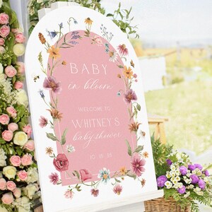 Op de afbeelding: Een wit, boogvormig bord met een roze midden, versierd met een bloemenkrans. Op het bord staat "BABY in bloom" en "WELCOME TO WHITNEY'S baby shower" in elegant schrift. De datum "10 15 26" staat onderaan. Een mand met bloemen staat op de voorgrond.