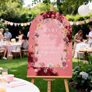 Op de afbeelding: Een roze bord op een houten ezel met de tekst "BABY in bloom" en "WELCOME TO WHITNEY'S baby shower". Het bord is versierd met bloemenillustraties en wordt opgesteld op een babyborrel in de buitenlucht.