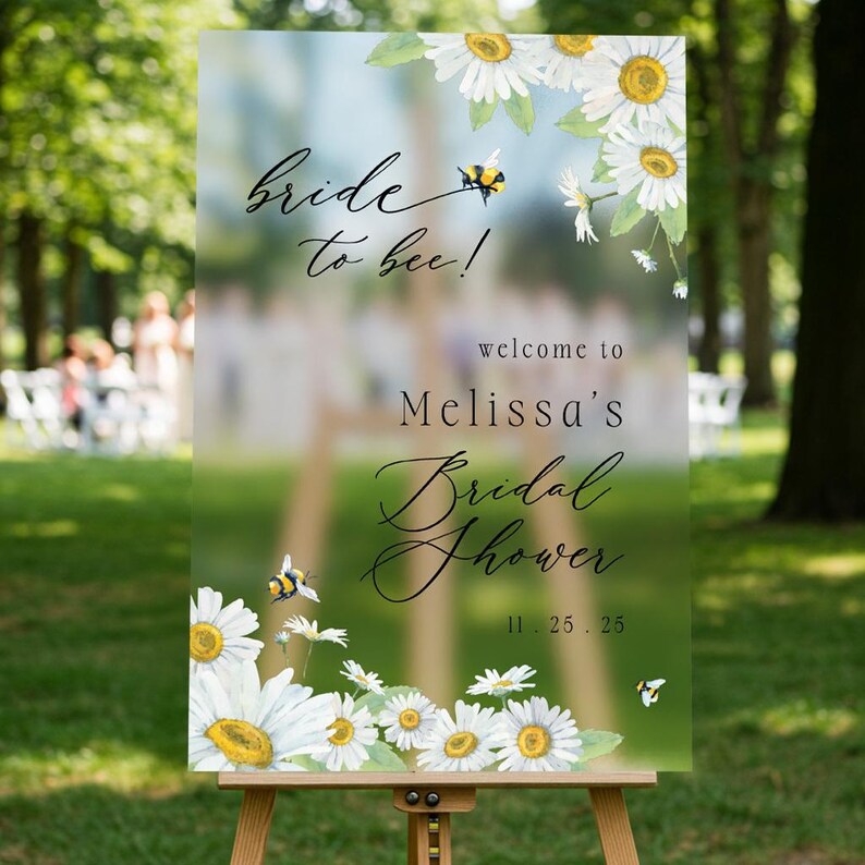 May include: A clear acrylic sign on an easel with the text "bride to bee!" and "welcome to Melissa's Bridal Shower 11.25.25". The sign is decorated with watercolor daisies and bees.