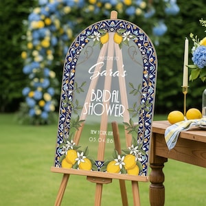 May include: A clear acrylic sign on a wooden easel, with the text "Welcome to Sara's Bridal Shower". The sign has a blue and white border with lemons and floral accents. The date and location are displayed. The easel is made of wood.